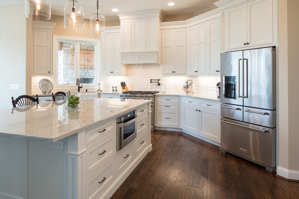 Large traditional u-shaped eat-in kitchen in Cincinnati with a farmhouse sink, shaker cabinets, white cabinets, marble benchtops, white splashback, glass tile splashback, stainless steel appliances, medium hardwood floors, with island and brown floor.