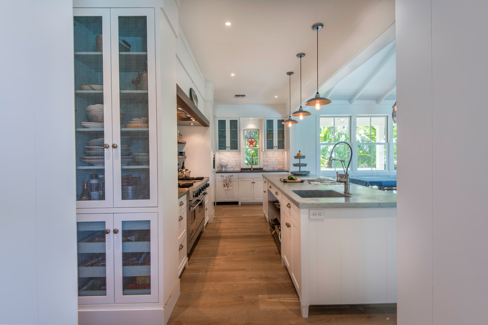 Photo of a mid-sized beach style l-shaped open plan kitchen in Tampa with shaker cabinets, white cabinets, limestone benchtops, white splashback, subway tile splashback, panelled appliances, with island, grey benchtop, an integrated sink, medium hardwood floors and brown floor.