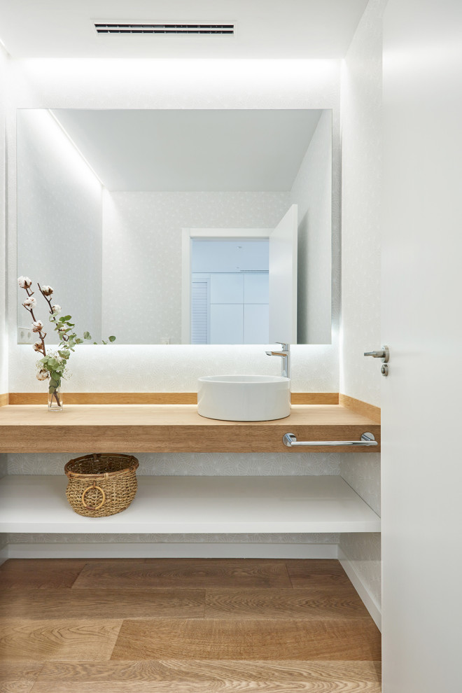 This is an example of a small contemporary powder room in Valencia with open cabinets, white cabinets, a one-piece toilet, yellow tile, white walls, medium hardwood floors, a vessel sink, wood benchtops, beige floor and beige benchtops.