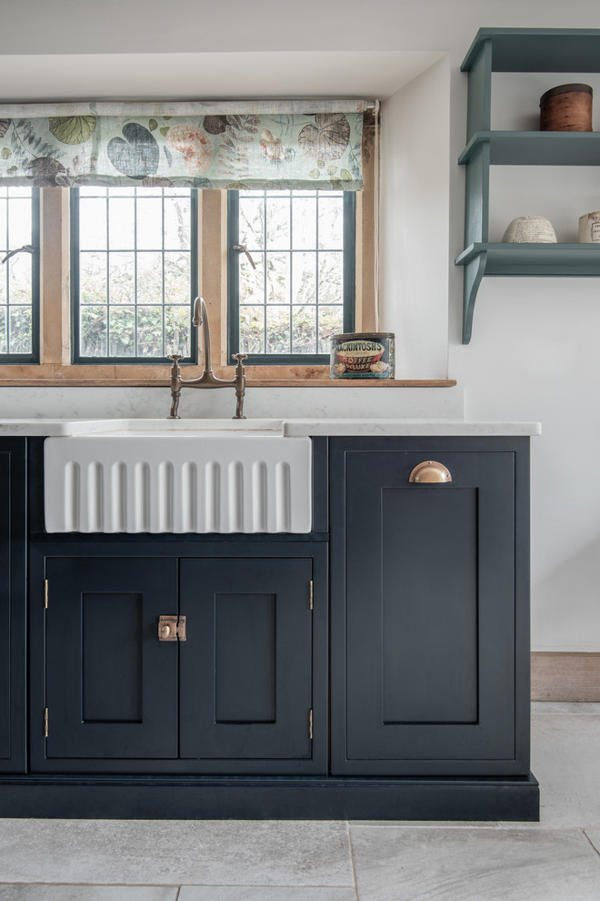 Mid-sized transitional u-shaped eat-in kitchen in Devon with a farmhouse sink, shaker cabinets, blue cabinets, solid surface benchtops, limestone floors, no island and white benchtop.