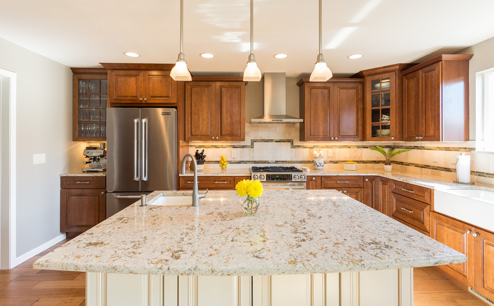 Inspiration for a large transitional l-shaped medium tone wood floor and brown floor eat-in kitchen remodel in Denver with a farmhouse sink, raised-panel cabinets, medium tone wood cabinets, granite countertops, beige backsplash, stone tile backsplash, stainless steel appliances and an island