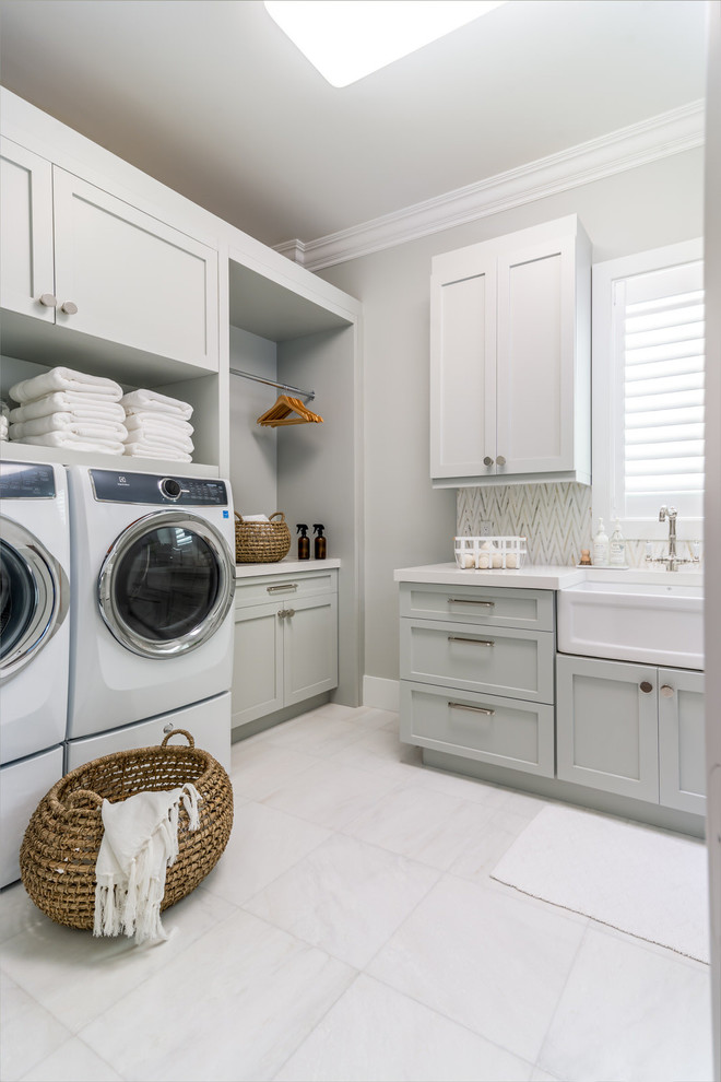 Inspiration for a transitional l-shaped dedicated laundry room in Miami with a farmhouse sink, shaker cabinets, grey cabinets, grey walls, a side-by-side washer and dryer, grey floor and white benchtop.