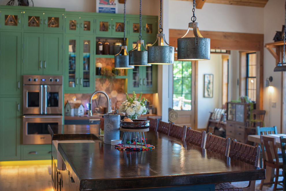 Example of a mid-sized mountain style l-shaped medium tone wood floor, brown floor and exposed beam open concept kitchen design in Seattle with a farmhouse sink, recessed-panel cabinets, green cabinets, soapstone countertops, brown backsplash, porcelain backsplash, stainless steel appliances, an island and black countertops