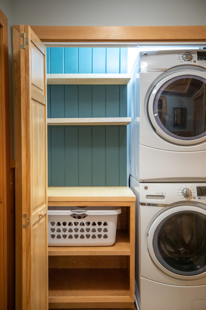 Photo of a small country single-wall laundry cupboard in Portland Maine with open cabinets, blue splashback, timber splashback, a stacked washer and dryer and green walls.