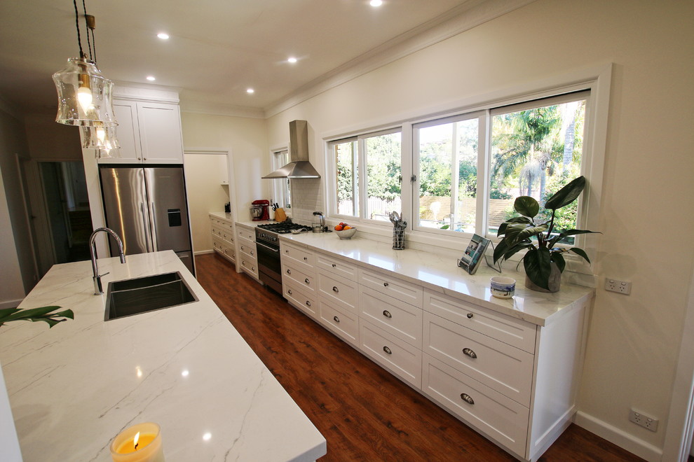 Photo of a large transitional galley open plan kitchen in Sydney with a double-bowl sink, shaker cabinets, white cabinets, quartz benchtops, white splashback, subway tile splashback, stainless steel appliances, medium hardwood floors, with island, brown floor and white benchtop.