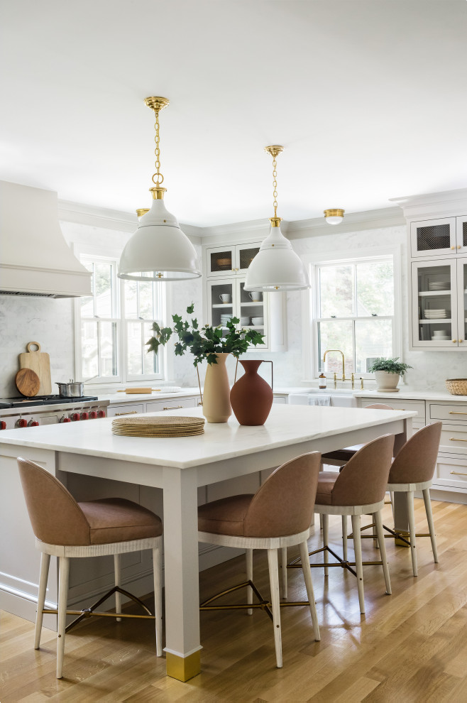 Traditional kitchen in Boston with a farmhouse sink, glass-front cabinets, beige cabinets, marble benchtops, white splashback, marble splashback, stainless steel appliances, light hardwood floors, with island and white benchtop.