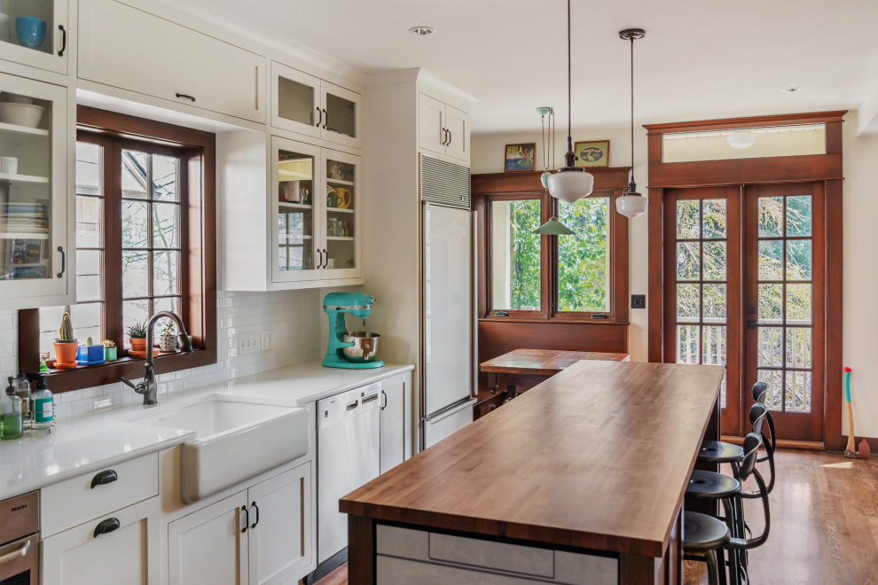 Inspiration for a mid-sized arts and crafts l-shaped eat-in kitchen in Seattle with a farmhouse sink, glass-front cabinets, marble benchtops, subway tile splashback, stainless steel appliances, dark hardwood floors, with island, white benchtop, white cabinets and white splashback.