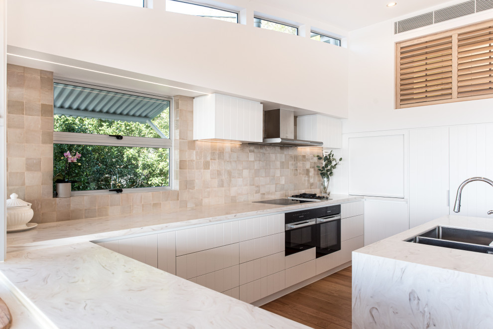Photo of a large beach style l-shaped eat-in kitchen in Sunshine Coast with a drop-in sink, white cabinets, beige splashback, porcelain splashback, black appliances, medium hardwood floors, with island and white benchtop.