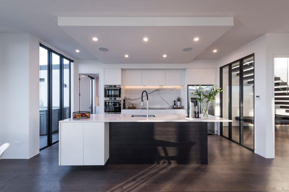 Large contemporary galley kitchen in Christchurch with an undermount sink, flat-panel cabinets, grey splashback, stainless steel appliances, with island, brown floor and white benchtop.