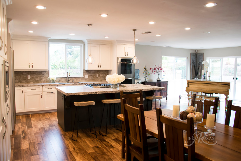 Large transitional l-shaped eat-in kitchen in San Diego with a single-bowl sink, shaker cabinets, white cabinets, granite benchtops, grey splashback, stone tile splashback, stainless steel appliances, medium hardwood floors, with island and brown floor.
