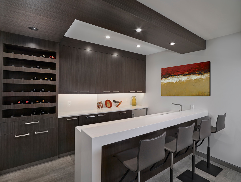Inspiration for a large contemporary galley seated home bar in Edmonton with flat-panel cabinets, dark wood cabinets, white splashback, grey floor, an undermount sink, solid surface benchtops, porcelain floors and white benchtop.