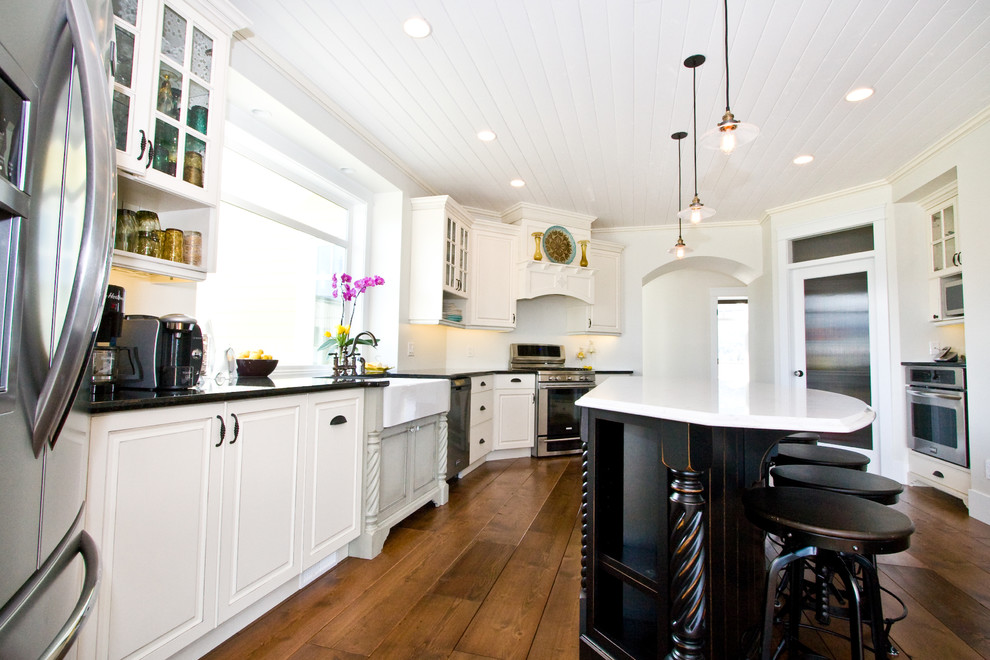 Design ideas for a large traditional u-shaped open plan kitchen in Vancouver with a farmhouse sink, raised-panel cabinets, white cabinets, quartz benchtops, stainless steel appliances, medium hardwood floors, with island, brown floor and white benchtop.