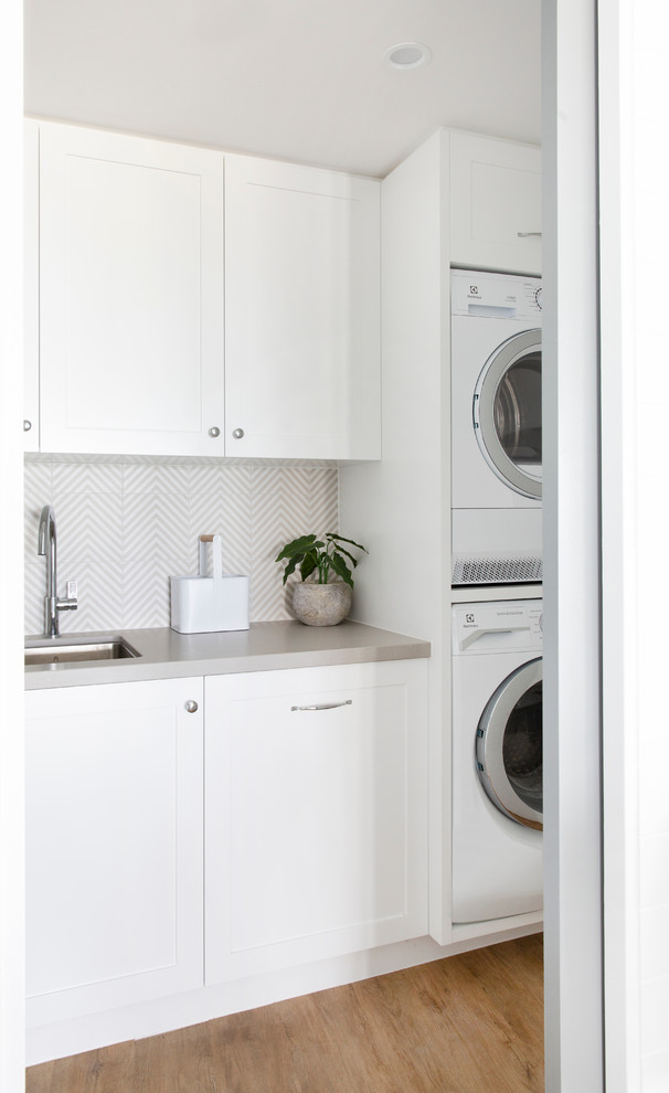 This is an example of a small beach style l-shaped utility room in Gold Coast - Tweed with an undermount sink, shaker cabinets, white cabinets, quartz benchtops, white walls, vinyl floors, a stacked washer and dryer, brown floor and grey benchtop.