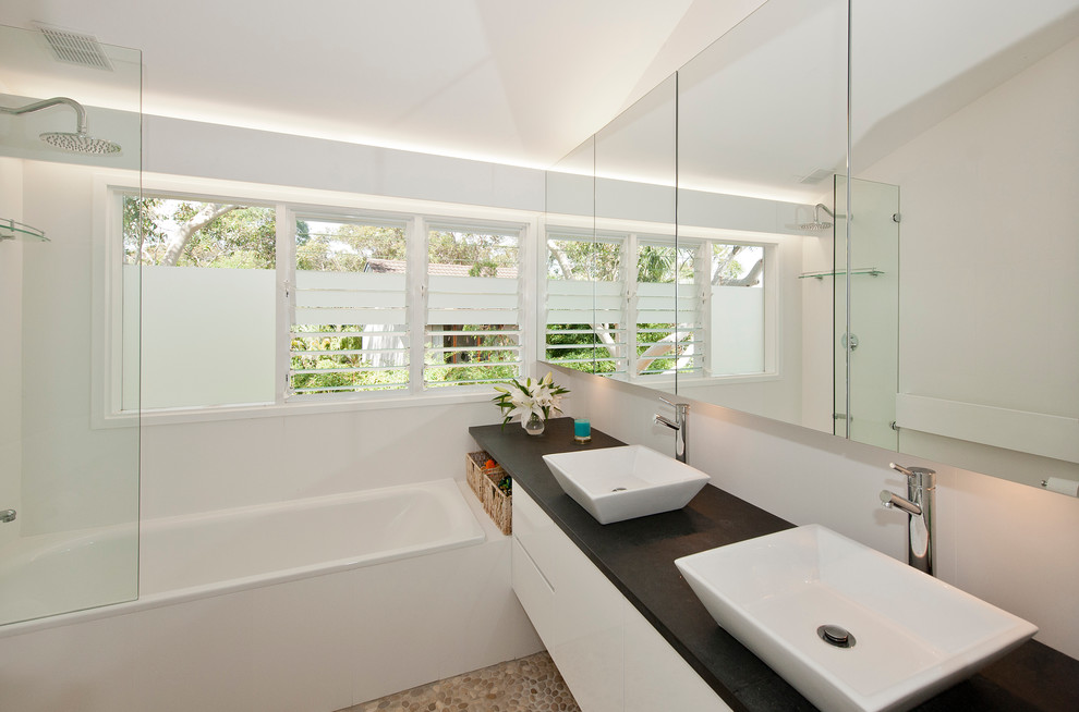 Inspiration for a mid-sized contemporary master bathroom in Sydney with a vessel sink, flat-panel cabinets, white cabinets, solid surface benchtops, a drop-in tub, a shower/bathtub combo, beige tile, white walls and pebble tile floors.