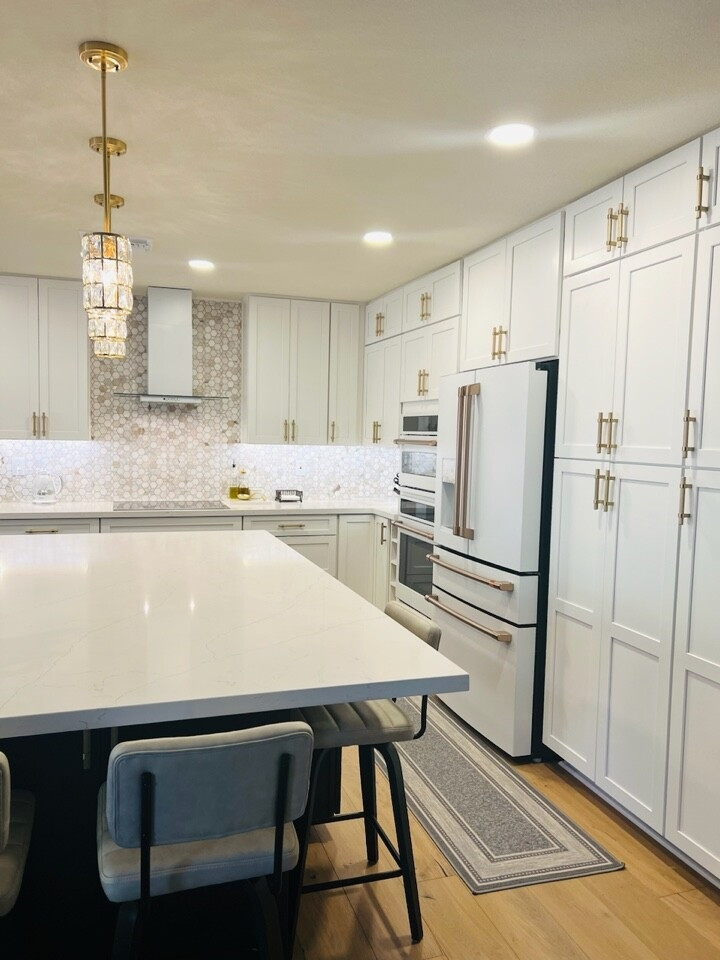Shabby chic-inspirerad inredning av ett mycket stort vit vitt kök, med en undermonterad diskho, skåp i shakerstil, vita skåp, bänkskiva i kvarts, beige stänkskydd, stänkskydd i mosaik, vita vitvaror, mellanmörkt trägolv och en köksö
