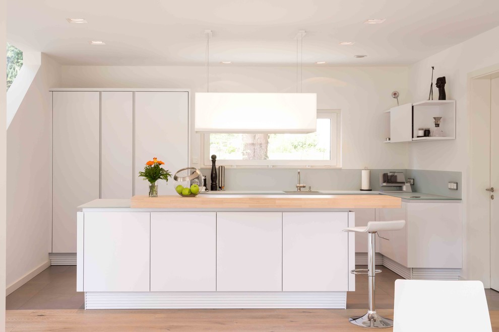 Mid-sized modern l-shaped open plan kitchen in Other with white cabinets, grey splashback, light hardwood floors, with island, brown floor, grey benchtop, an integrated sink, flat-panel cabinets and glass sheet splashback.