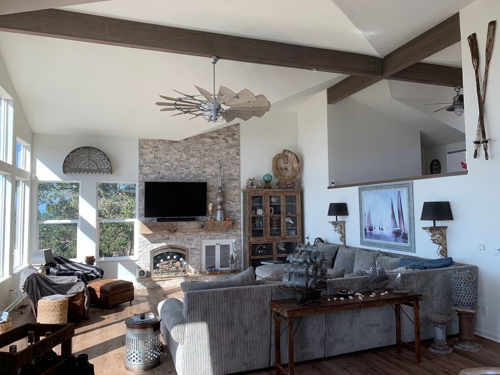 This is an example of a large beach style enclosed family room in San Luis Obispo with white walls, medium hardwood floors, a standard fireplace, a stone fireplace surround, a wall-mounted tv and brown floor.