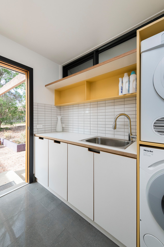 Contemporary single-wall utility room in Canberra - Queanbeyan with a drop-in sink, flat-panel cabinets, white cabinets, white walls, concrete floors, a stacked washer and dryer, grey floor and white benchtop.