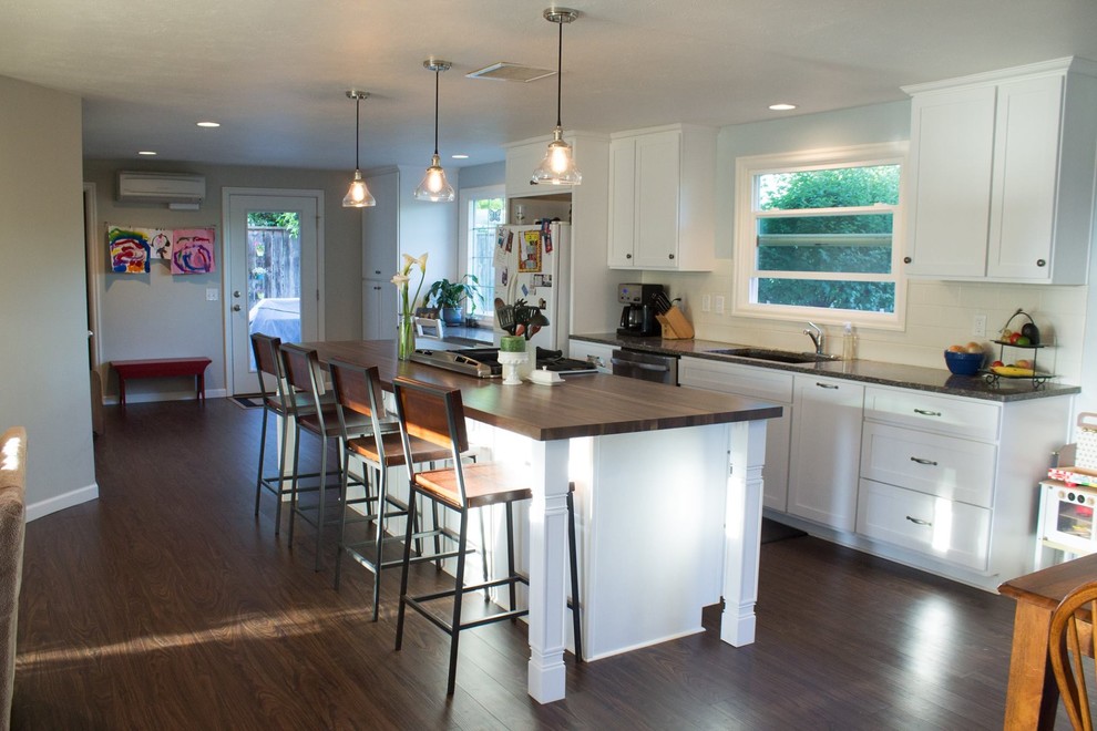 Example of a mid-sized transitional galley dark wood floor and brown floor open concept kitchen design with an undermount sink, shaker cabinets, white cabinets, wood countertops, white backsplash, porcelain backsplash, white appliances and an island