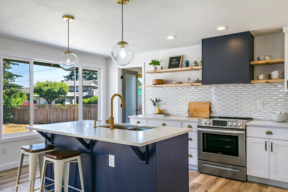 This is an example of a large midcentury l-shaped kitchen in Seattle with a drop-in sink, shaker cabinets, white cabinets, white splashback, stainless steel appliances, laminate floors, with island, brown floor, white benchtop, quartz benchtops and ceramic splashback.