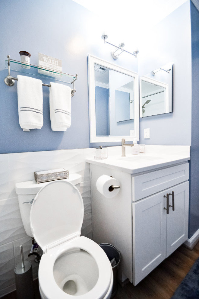 Photo of a small beach style bathroom in DC Metro with shaker cabinets, white cabinets, a drop-in tub, a shower/bathtub combo, a two-piece toilet, white tile, ceramic tile, blue walls, wood-look tile, an integrated sink, engineered quartz benchtops, brown floor, a shower curtain, white benchtops, a niche, a single vanity and a built-in vanity.