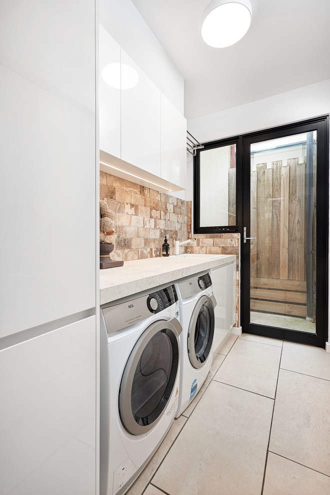 Photo of a single-wall laundry room in Melbourne with a drop-in sink, white cabinets, solid surface benchtops, ceramic floors, a side-by-side washer and dryer, beige floor and beige benchtop.