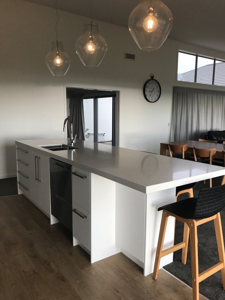 Design ideas for a large contemporary galley open plan kitchen in Christchurch with a single-bowl sink, flat-panel cabinets, white cabinets, quartz benchtops, white splashback, glass sheet splashback, stainless steel appliances, vinyl floors, with island, brown floor and white benchtop.