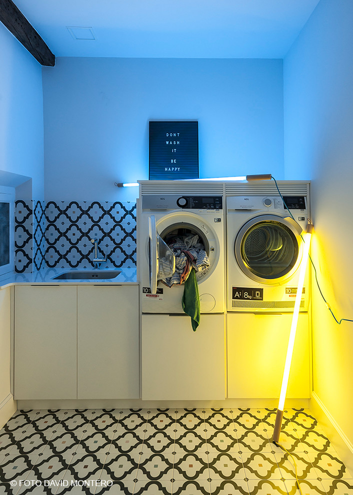Photo of a small eclectic single-wall dedicated laundry room in Other with an undermount sink, ceramic floors, flat-panel cabinets, white cabinets, quartzite benchtops, black walls, a stacked washer and dryer and multi-coloured floor.