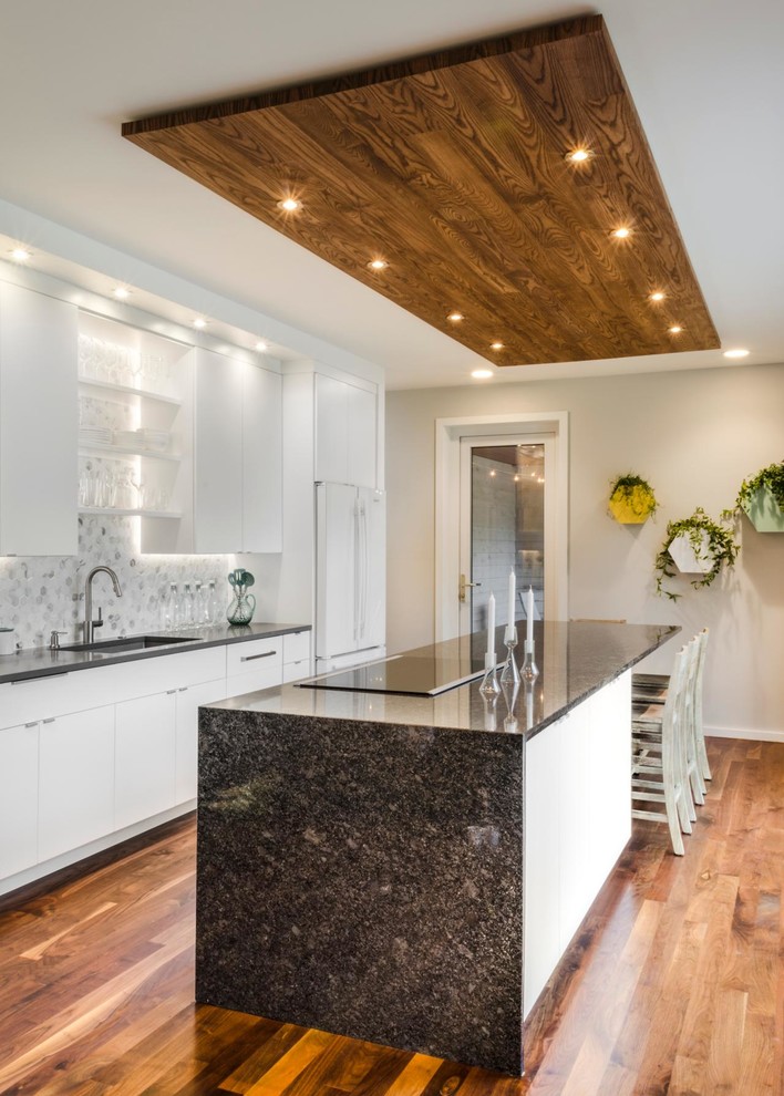 Inspiration for a mid-sized contemporary single-wall open plan kitchen in Toronto with an undermount sink, flat-panel cabinets, white cabinets, grey splashback, marble splashback, white appliances, medium hardwood floors, with island, brown floor and brown benchtop.