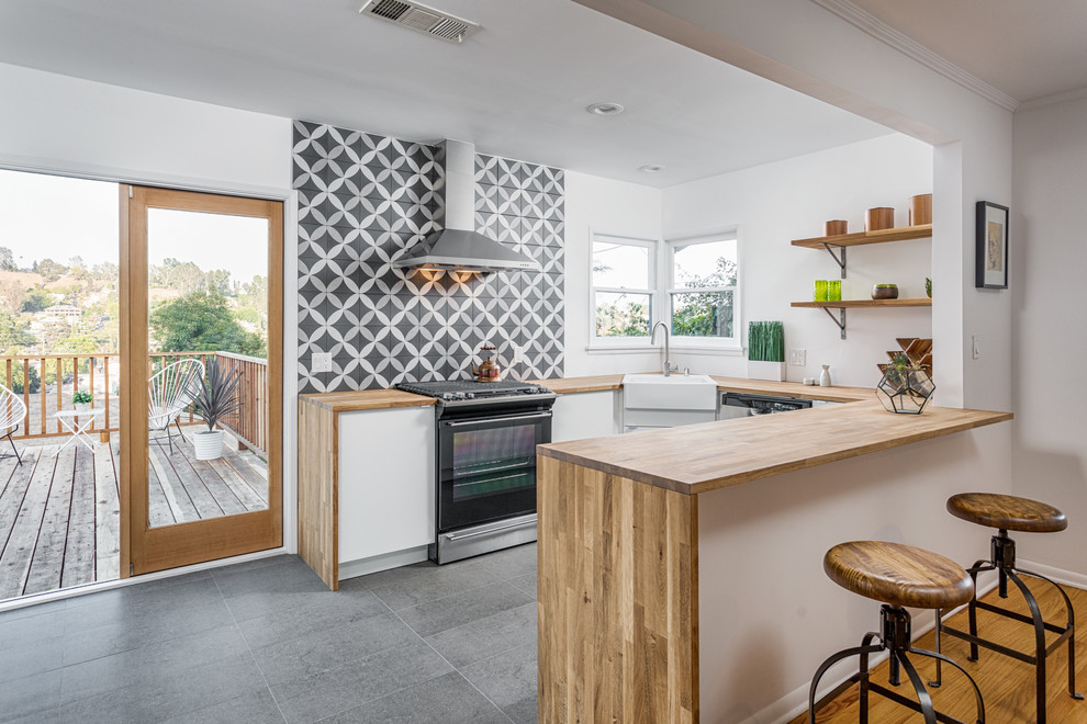 Inspiration for a mid-sized contemporary u-shaped open plan kitchen in Los Angeles with a farmhouse sink, wood benchtops, multi-coloured splashback, black appliances, a peninsula, flat-panel cabinets, medium wood cabinets, ceramic splashback, slate floors and beige benchtop.