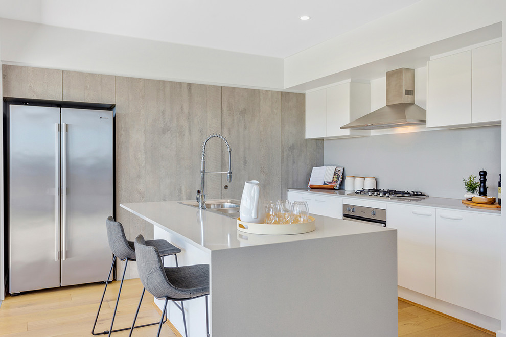 Inspiration for a contemporary l-shaped kitchen in Brisbane with a drop-in sink, flat-panel cabinets, white cabinets, grey splashback, stainless steel appliances, light hardwood floors, with island, beige floor and white benchtop.