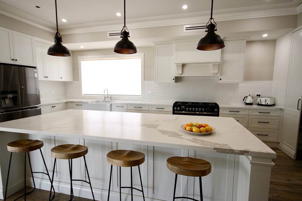 Design ideas for a large traditional l-shaped eat-in kitchen in Sydney with a farmhouse sink, shaker cabinets, white cabinets, quartz benchtops, white splashback, ceramic splashback, black appliances, medium hardwood floors, with island, brown floor and white benchtop.