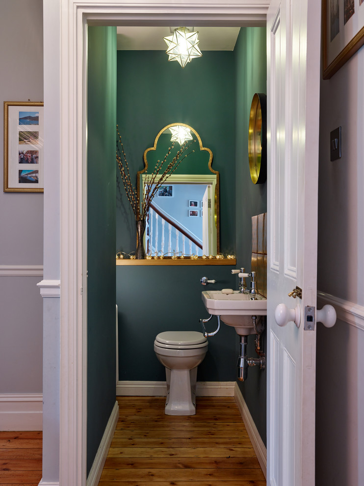 Photo of a small transitional powder room in Other with a one-piece toilet, green walls, medium hardwood floors, a wall-mount sink and brown floor.