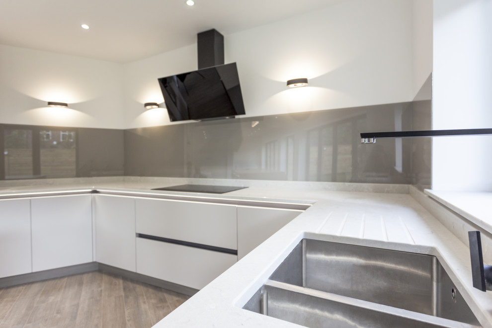 Photo of a mid-sized contemporary u-shaped eat-in kitchen in Manchester with an undermount sink, flat-panel cabinets, grey cabinets, quartzite benchtops, brown splashback, glass sheet splashback, black appliances, medium hardwood floors, with island, brown floor and grey benchtop.