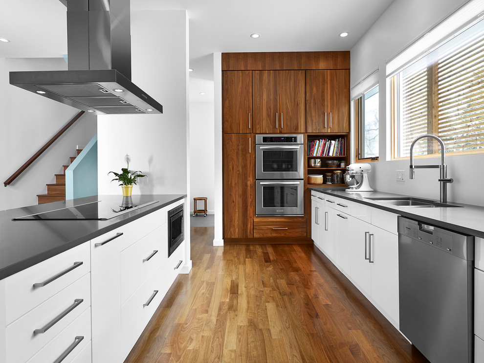 This is an example of a mid-sized modern galley open plan kitchen in Edmonton with flat-panel cabinets, stainless steel appliances, an undermount sink, white cabinets, solid surface benchtops, medium hardwood floors, a peninsula and brown floor.
