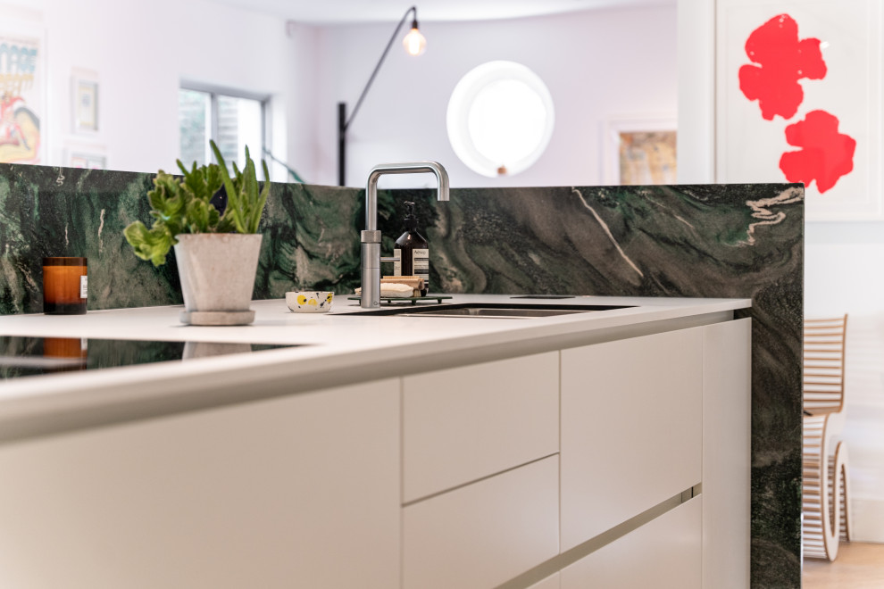 This is an example of a medium sized modern galley open plan kitchen in London with a built-in sink, flat-panel cabinets, white cabinets, quartz worktops, green splashback, granite splashback, black appliances, light hardwood flooring, an island, brown floors, white worktops and a drop ceiling.