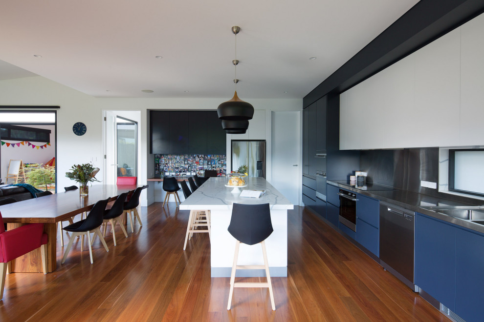 Contemporary l-shaped open plan kitchen in Sydney with flat-panel cabinets, blue cabinets, stainless steel benchtops, metallic splashback, medium hardwood floors, with island, white benchtop, an integrated sink, metal splashback, stainless steel appliances and brown floor.