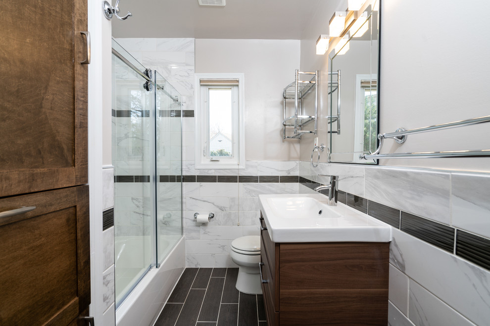 Photo of a mid-sized contemporary 3/4 bathroom in DC Metro with a shower/bathtub combo, a one-piece toilet, an integrated sink, a sliding shower screen, dark wood cabinets, an alcove tub, gray tile, grey walls, black floor, white benchtops, marble, vinyl floors, engineered quartz benchtops and shaker cabinets.