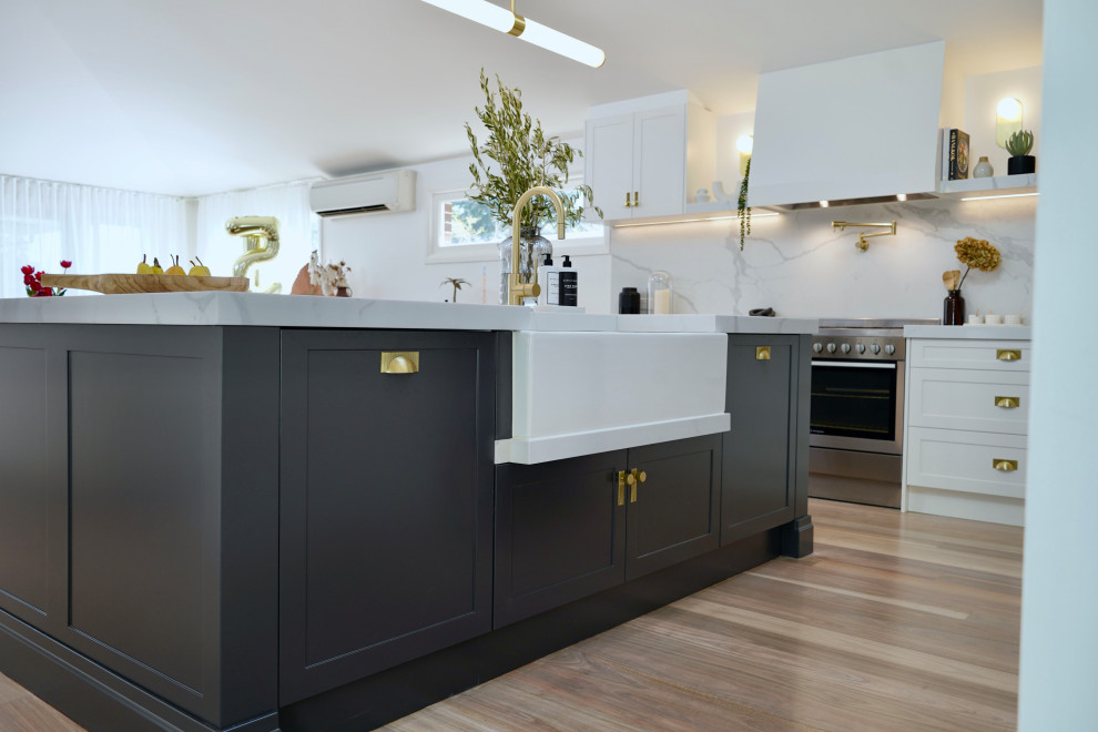 This is an example of a large classic l-shaped open plan kitchen in Sydney with a belfast sink, shaker cabinets, white cabinets, engineered stone countertops, white splashback, engineered quartz splashback, stainless steel appliances, light hardwood flooring, an island, brown floors and white worktops.