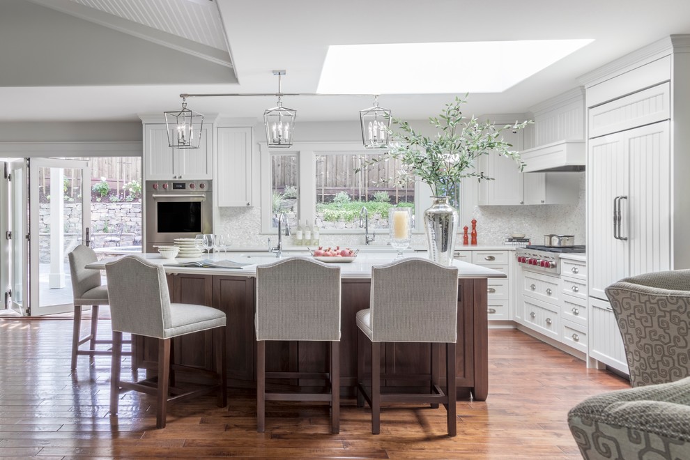 This is an example of a large traditional l-shaped open plan kitchen in San Francisco with a farmhouse sink, shaker cabinets, white cabinets, quartz benchtops, white splashback, mosaic tile splashback, panelled appliances, medium hardwood floors, with island and brown floor.