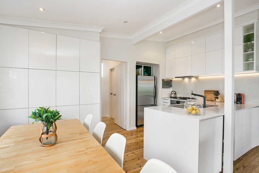 Beach style u-shaped kitchen in Central Coast with a double-bowl sink, flat-panel cabinets, white cabinets, white splashback, stainless steel appliances, medium hardwood floors, a peninsula, brown floor and white benchtop.
