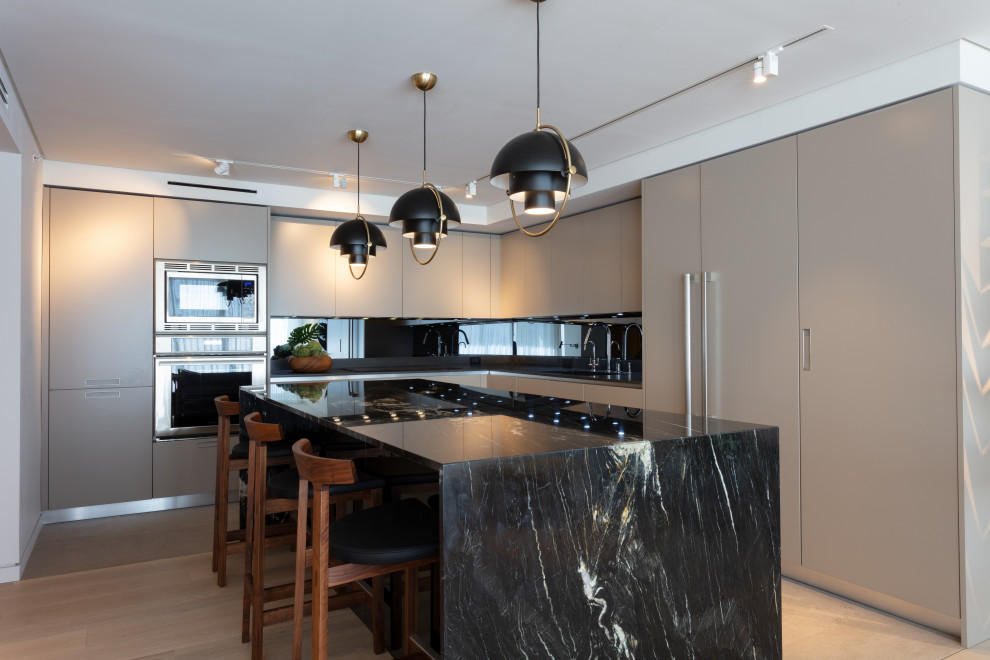 Design ideas for a contemporary l-shaped kitchen in Miami with beige cabinets, marble benchtops, black splashback, stainless steel appliances, light hardwood floors, with island, black benchtop, flat-panel cabinets, mirror splashback and beige floor.