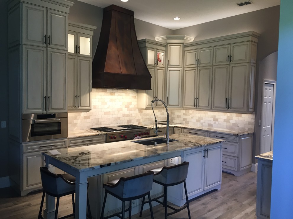 Design ideas for a mid-sized transitional kitchen in Orlando with a triple-bowl sink, raised-panel cabinets, distressed cabinets, granite benchtops, beige splashback, stone tile splashback, stainless steel appliances, porcelain floors and with island.