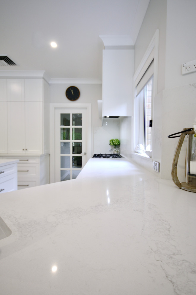 This is an example of a large transitional open plan kitchen in Sydney with a farmhouse sink, recessed-panel cabinets, white cabinets, quartz benchtops, white splashback, engineered quartz splashback, stainless steel appliances, ceramic floors, with island, grey floor and white benchtop.
