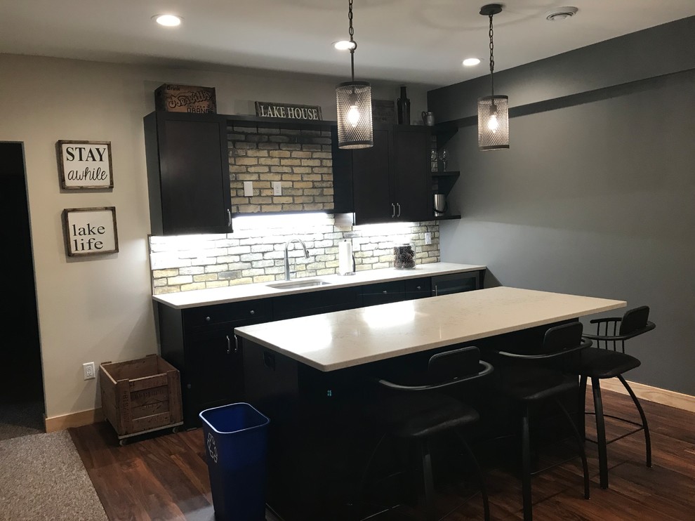 This is an example of a country single-wall seated home bar in Milwaukee with an undermount sink, shaker cabinets, dark wood cabinets, quartz benchtops, beige splashback, brick splashback, vinyl floors, brown floor and beige benchtop.