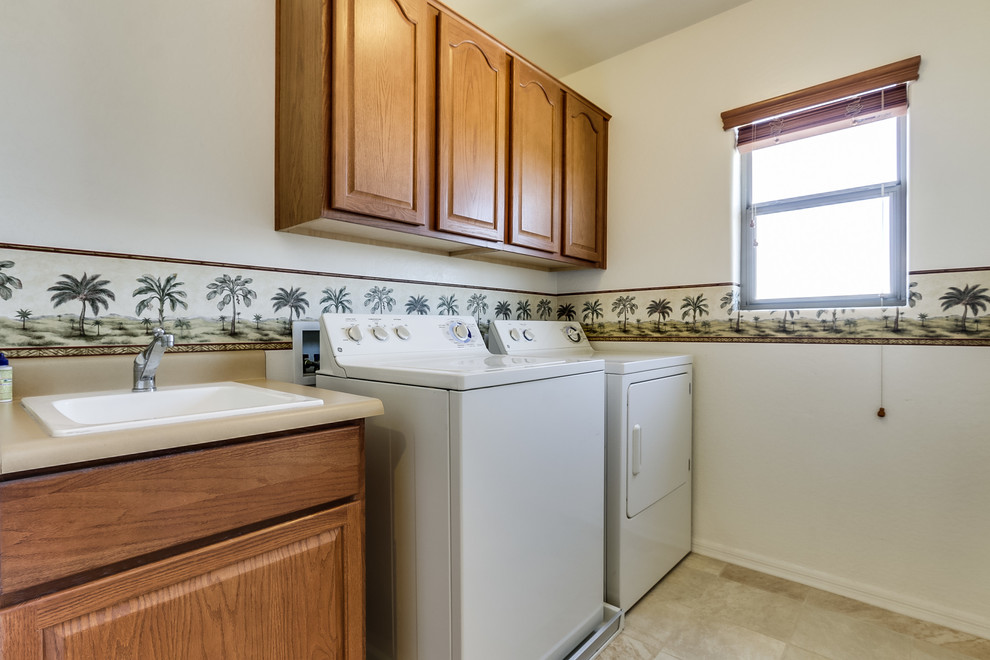 Inspiration for a large traditional galley dedicated laundry room in Phoenix with a drop-in sink, raised-panel cabinets, medium wood cabinets, solid surface benchtops, white walls, travertine floors and a side-by-side washer and dryer.