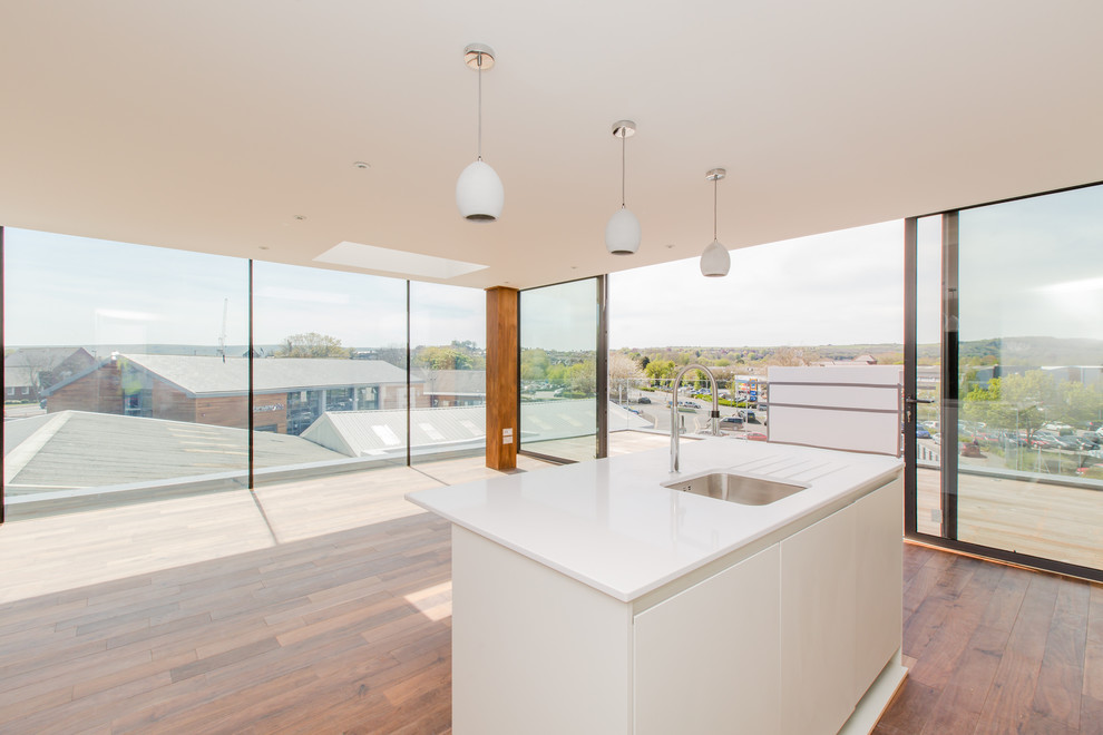 Mid-sized industrial l-shaped open plan kitchen in Sussex with a drop-in sink, flat-panel cabinets, white cabinets, quartz benchtops, glass sheet splashback, panelled appliances, medium hardwood floors and with island.