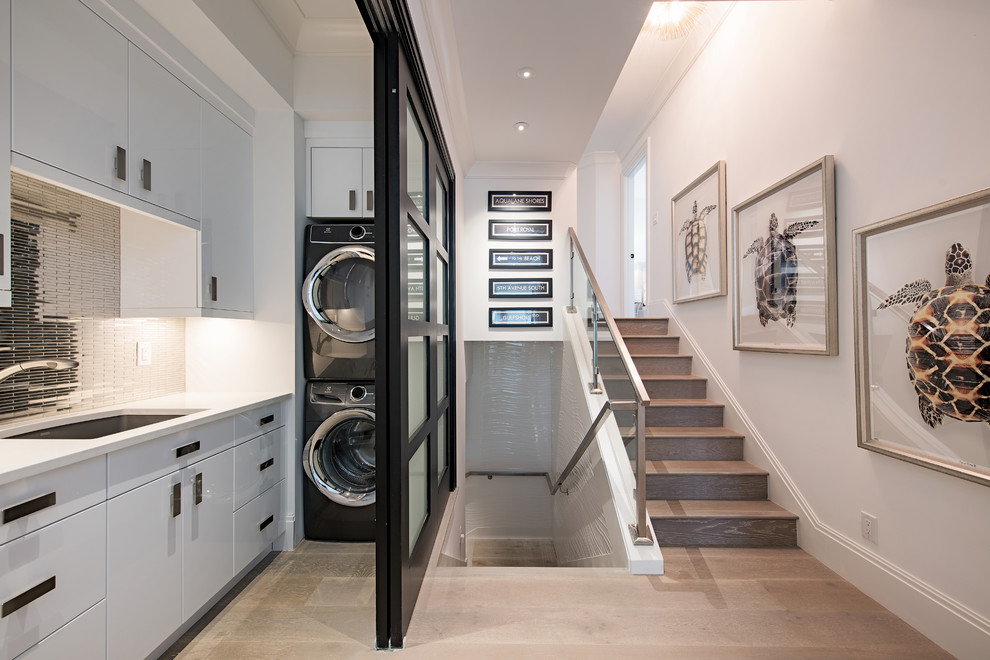 Photo of a transitional l-shaped dedicated laundry room in Miami with a single-bowl sink, flat-panel cabinets, white cabinets, white walls, light hardwood floors, a stacked washer and dryer, beige floor and white benchtop.
