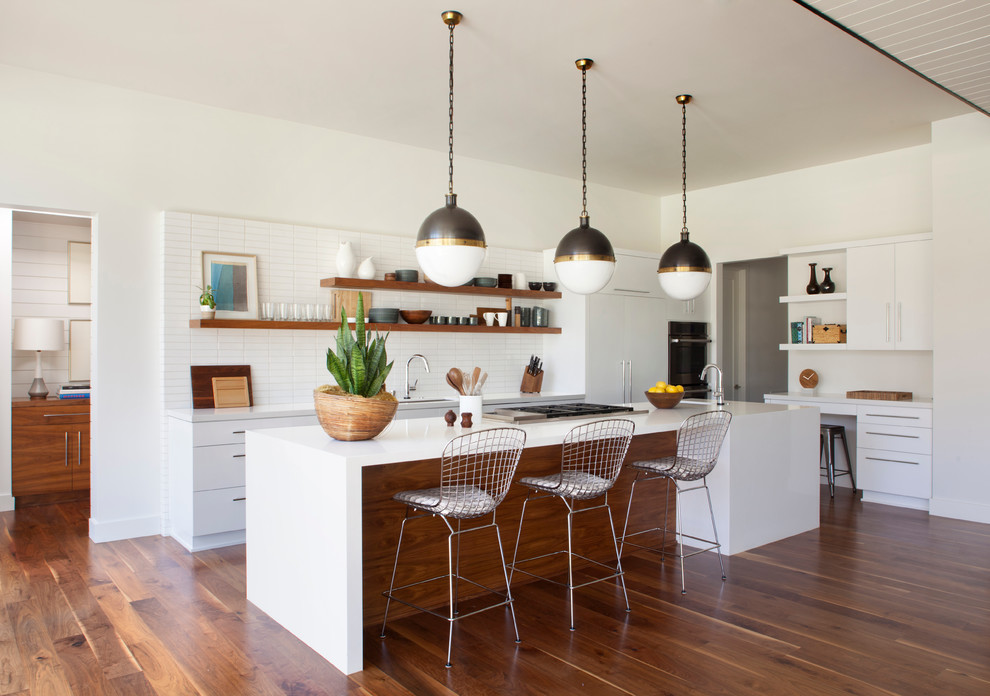 Design ideas for a mid-sized midcentury l-shaped open plan kitchen in Los Angeles with flat-panel cabinets, white cabinets, quartz benchtops, white splashback, ceramic splashback, panelled appliances, medium hardwood floors and with island.