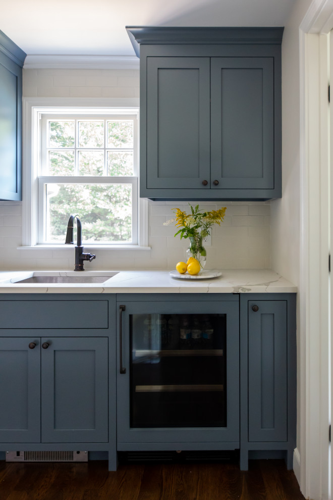 Foto på ett vintage vit kök, med en nedsänkt diskho, luckor med profilerade fronter, blå skåp, marmorbänkskiva, vitt stänkskydd, stänkskydd i tunnelbanekakel, integrerade vitvaror, mellanmörkt trägolv, en köksö och brunt golv